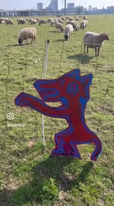 Painted wolf in a field with sheep – urban art by Art Is Trash Painted wolf figure on a board in a field among sheep, Art Is Trash